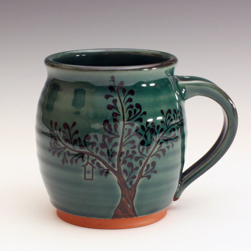 Red earthenware mug with green glaze. The trunk is carved into the clay and black decoration for the leaves. Some contain bird houses carved in. The roots are carved into the base of the mug.