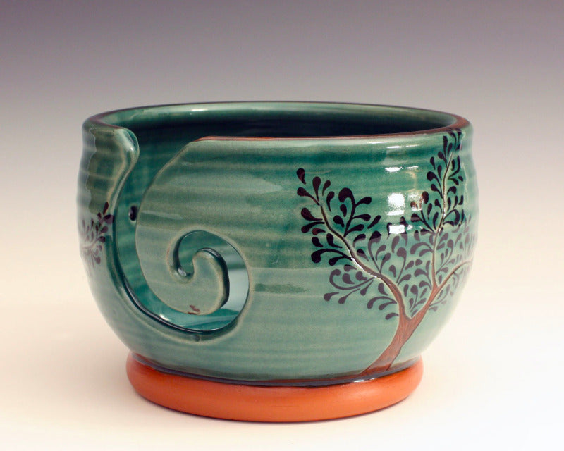 Ceramic yarn bowl.  Glazed green with trees decorating the outside.  Swirl cut out of the side as well as three holes.  