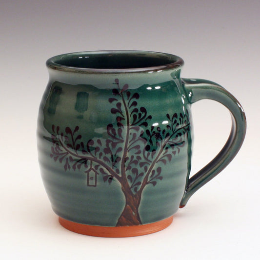 Red earthenware mug with green glaze.  The trunk is carved into the clay and black decoration for the leaves.  Some contain bird houses carved in.  The roots are carved into the base of the mug.  